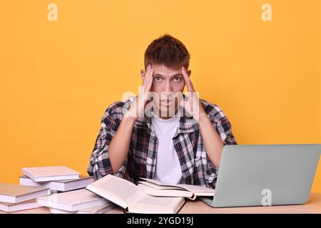 Young student with books and laptop having stress before exam at table against yellow background Stock Photo