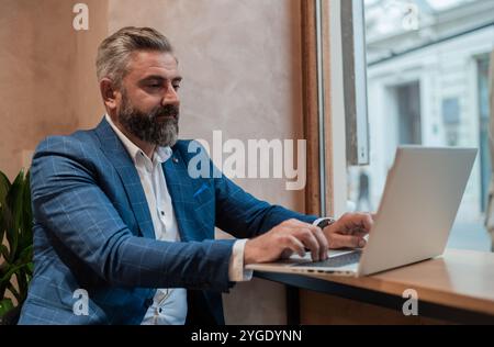 Portrait of a mature business man sitting at his desk in the office or restaurant cafe near the window and working on a laptop. Stock Photo