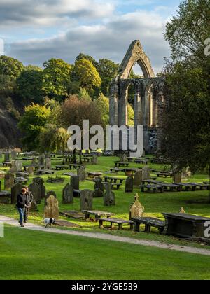 Picturesque scenic sunlit monastic Bolton Abbey ruins (gravestones & graves, couple of dog walkers, beautiful setting) - Yorkshire Dales, England, UK. Stock Photo