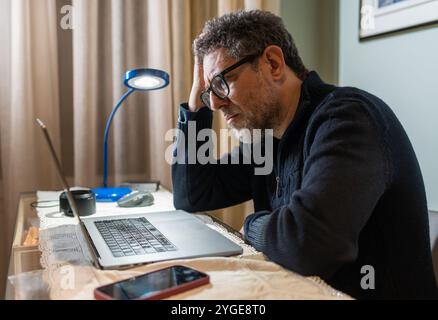 Exhausted middle aged businessman feeling stressed and overwhelmed while working on laptop computer at home office desk Stock Photo