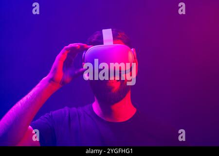 Man with VR (virtual reality) goggles touching and adjusting the headset. Wearing a device for multimedia and gaming. Colorful studio light. Stock Photo