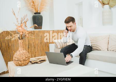 A young man sits at home on the sofa and works remotely on a laptop. Stock Photo