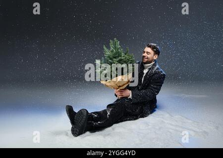 The young man smiles as he holds a festive tree while surrounded by falling snowflakes. Stock Photo