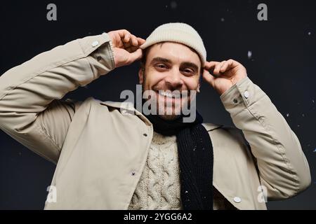 A cheerful young man smiles broadly while adjusting his warm hat in a winter setting with snow. Stock Photo