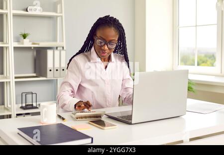 Young African American woman accountant sitting in office, working on laptop and using calculator Stock Photo
