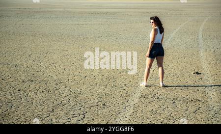 Young woman in shorts and sunglasses stands on dry desert ground. Stock Photo