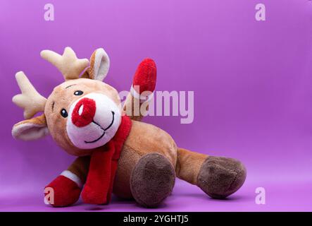 A stuffed reindeer is laying on a purple background. The reindeer is wearing a red scarf and has a red nose Stock Photo