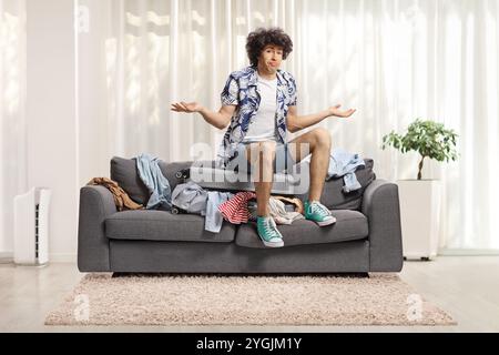 Confused young man sitting on top of an overpacked suitcase on a sofa at home Stock Photo
