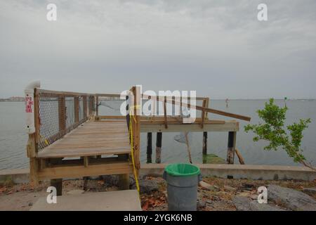 POV leading lines view damaged Wooden boardwalk. Trash can,Green trees on the side. Damaged seawall and sidewalk. Gulfport Florida Beach. On Boca Cieg Stock Photo