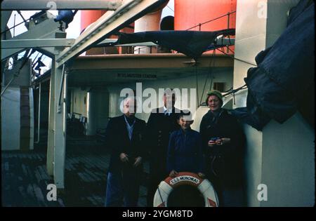 Group Posing with TSS Monowai Life Ring on Deck During Final Voyage: A rare color photograph from the final voyage of the TSS Monowai, capturing a group of people, including a child, standing on the deck near a life-saving ring labeled “TSS Monowai.” The ship’s iconic red funnels are prominently visible in the background. This historic voyage marked the end of an era for the trans-Tasman passenger liner, which had been a significant vessel connecting New Zealand and Australia. Stock Photo