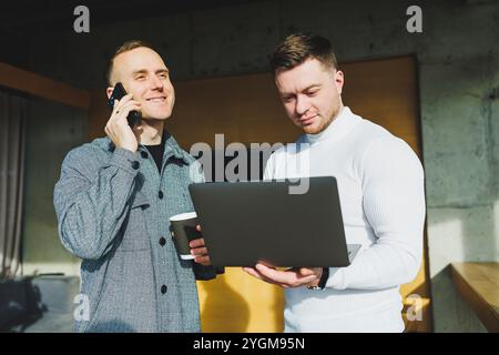 Work of two male colleagues standing in office together while exchanging ideas with laptop in meeting room and analyzing work schemes. Work in a moder Stock Photo
