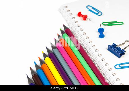 A variety of colorful pencils are arranged alongside paperclips, push pins, and a binder clip on a spiral notebook Stock Photo