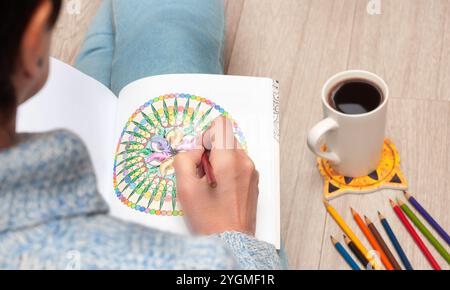 A person relaxing while coloring a mandala in an adult coloring book, accompanied by colored pencils and a cup of coffee for a peaceful experience. Stock Photo