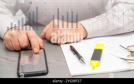 A person reviews cryptocurrency data on a smartphone and makes notes while analyzing market trends in a contemporary office environment. Stock Photo