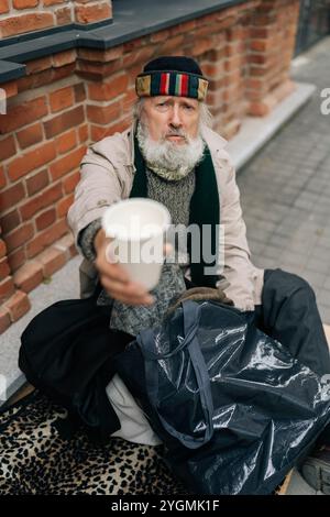 Vertical portrait of aged homeless man sits on urban street with belongings, seeking assistance, holding disposable cup and begging for alms. Stock Photo