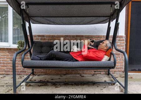 A man in red is lying on a swinging sofa in the garden of his house with his cat. Relaxed, comfortable, stress-free, free time, not working, resting Stock Photo