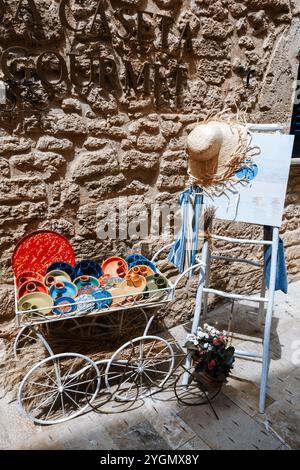 Alcudia Mallorca, Rustic Mediterranean decor: Colorful ceramic plates in vintage metal cart, straw hat on ladder, stone wall background. Traditional E Stock Photo