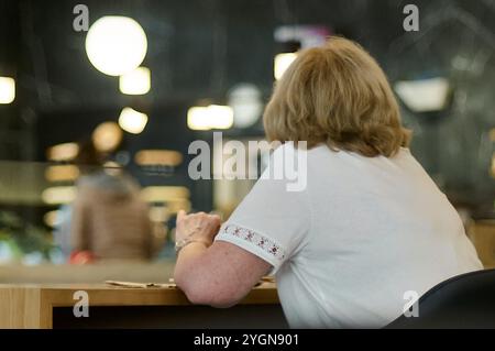 Viladecans. Spain - November 08,2024: Woman with short blonde hair, wearing a white shirt, seated at a wooden table in a contemporary coffee shop, wit Stock Photo