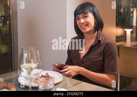 Young professional woman using smartphone during dinner at upscale restaurant, smiling while checking social media Stock Photo