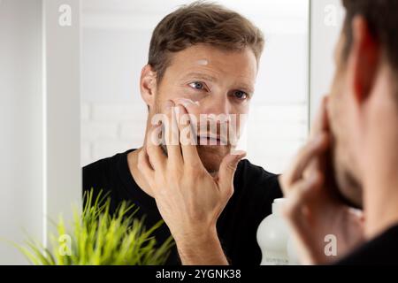 man applying facial cream in front of mirror at bathroom. male face skin care Stock Photo