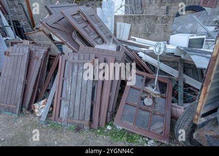 A pile of discarded wooden shutters, doors, and windows with cracked paint, rusted hinges, and glass panes, Stock Photo