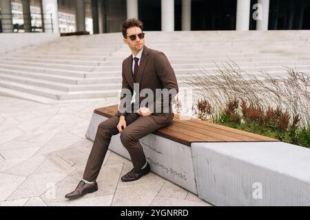 Full length portrait of of stylish photogenic young businessman in brown suit and sunglasses sitting confidently on city bench. Stock Photo