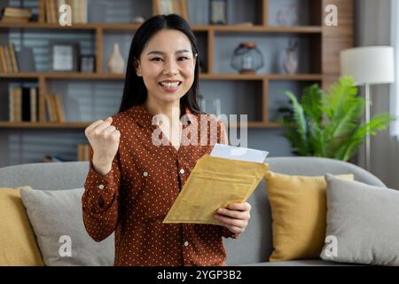 Asian woman expressing joy at home holding envelope in hand. Her happy and victorious expression represents success and positive news. Setting is cozy and comfortable. Stock Photo