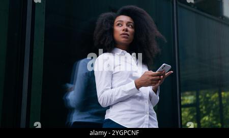 African American woman female girl thinking decision pondering urban city user mobile phone smartphone businesswoman business employee chat social Stock Photo