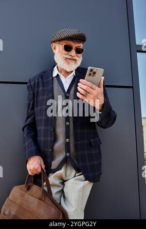 A well-dressed senior man with a beard stands casually, using his smartphone while enjoying the fall weather. Stock Photo