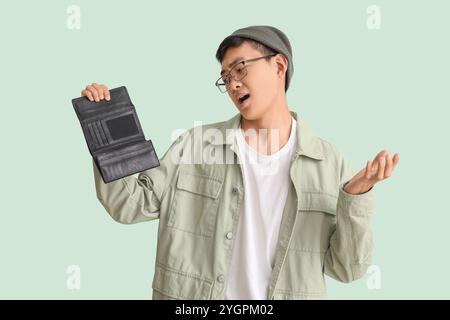 Upset young Asian man with empty wallet on green background Stock Photo