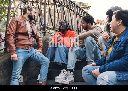 Diverse group of young friends laughing and talking outdoors in urban setting, authentic lifestyle meetup - casual young people meetup Stock Photo