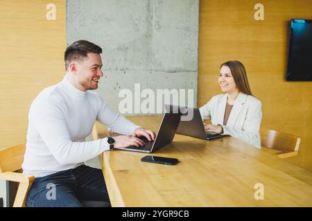 Male and female colleagues are working at a wooden table on laptops and talking. Concept of business cooperation and teamwork. Young colleagues in the Stock Photo