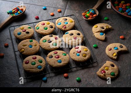 Vanilla cookies with red, blue, green and yellow chocolate chips on black grill, surrounded by chocolate chips and bitten cookies on ceramic table Stock Photo