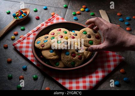 Man's hand taking a vanilla cookie with red, blue, green and yellow chocolate chips from a group of cookies on a round black plate on a red napkin wit Stock Photo