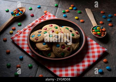 Group of red, blue, green and yellow vanilla chocolate chip cookies on a round black plate on a red napkin with white checks, surrounded by chocolate Stock Photo