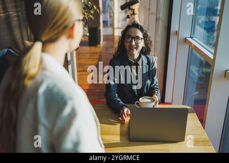 Positive female colleagues in casual clothes looking at netbook screen taking notes about project at table with coffee cups in office. Students work. Stock Photo