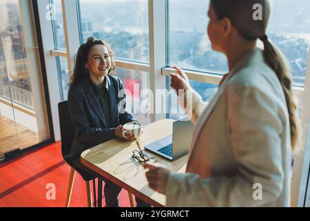 Positive female colleagues in casual clothes looking at netbook screen taking notes about project at table with coffee cups in office. Students work. Stock Photo