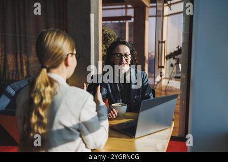 Positive female colleagues in casual clothes looking at netbook screen taking notes about project at table with coffee cups in office. Students work. Stock Photo