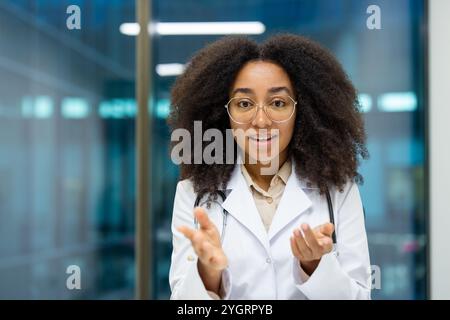 African American woman doctor wearing white coat in modern hospital, engaged in video call. Confident expression, looking at camera, conveying care, professionalism. Ideal for medical, technology concepts. Stock Photo