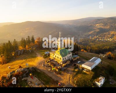 The Tanvaldsky Spicak lookout tower stands prominently in the Jizera Mountains, surrounded by vibrant autumn foliage during a serene evening glow. Visitors are seen enjoying the view. Stock Photo