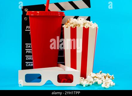 Image of movie clapperboard behind red soda glass, bucket of popcorn and 3D glasses on blue background, cinema entertainment concept Stock Photo