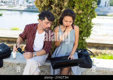 Two individuals engaged in a focused discussion over a laptop, sitting on a bench in a sunny park environment by the water. Stock Photo