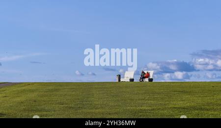 Panoramic photo of a grassy dyke with two white benches and two tourists sitting in the sun with blue sky in the background, Unesco World Heritage Wad Stock Photo