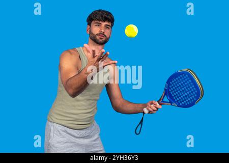 Young male athlete playing paddle tennis, about to hit the ball with his racket on a vibrant blue background Stock Photo