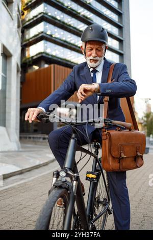 Senior commuter with helmet and bag checking time while traveling to work on bicycle in modern city Stock Photo