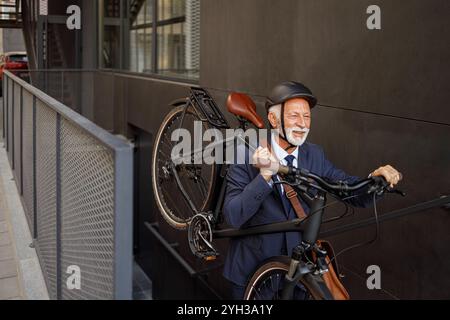 Positive senior male professional carrying his bicycle on shoulder and moving up on steps in city Stock Photo