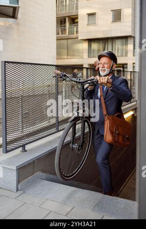 Smiling senior male manager with bag and helmet carrying his bicycle and walking up on steps in city Stock Photo