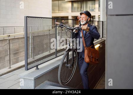 Senior male commuter in helmet carrying his bicycle on shoulder while walking on steps in modern city Stock Photo