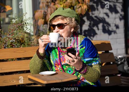 An eighty year old grandmother with a smartphone drinks coffee in a outdoor cafe on a sunny autumn day Stock Photo