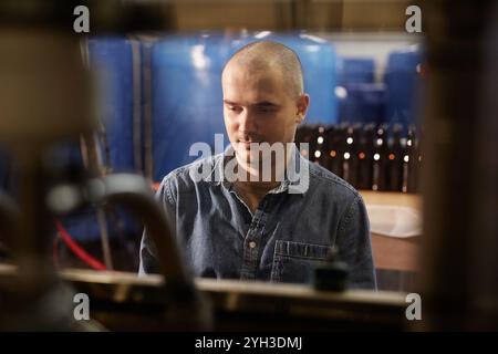 Shot through factory machinery of happy male production worker wearing denim overalls directing factory equipment on assembly line in workshop of cide Stock Photo